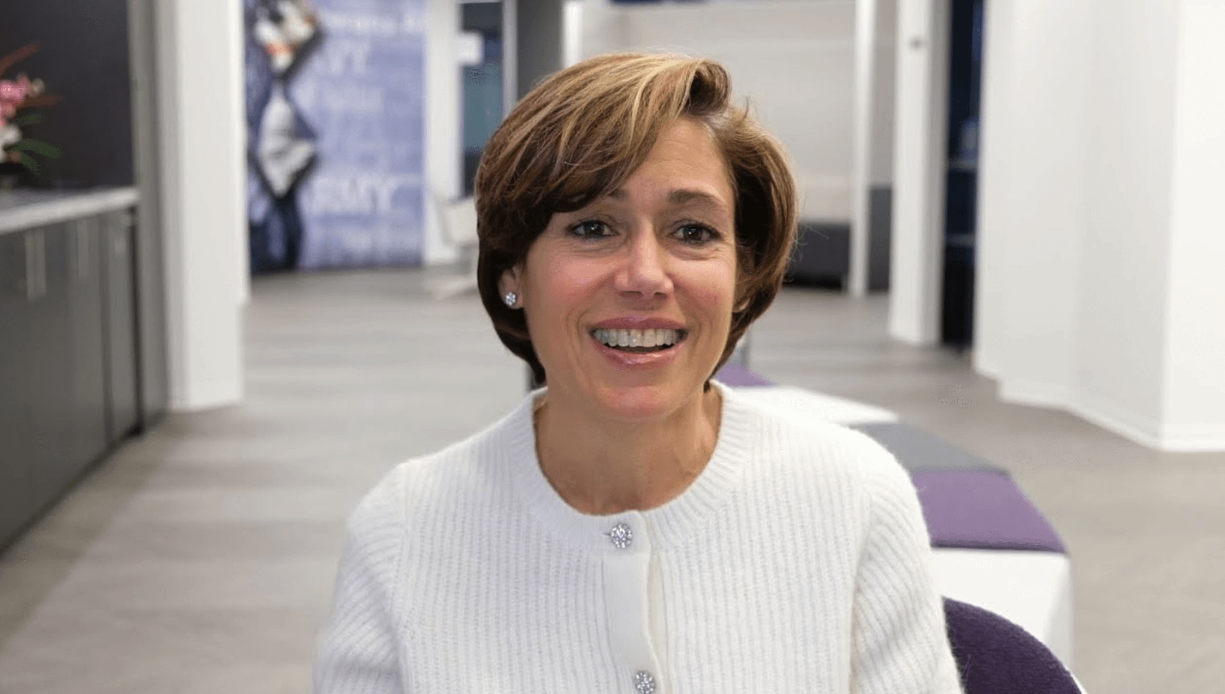 Gina Grosso smiling in a white cardigan, seated in a Human Resources office hallway with purple and gray seating in the background.