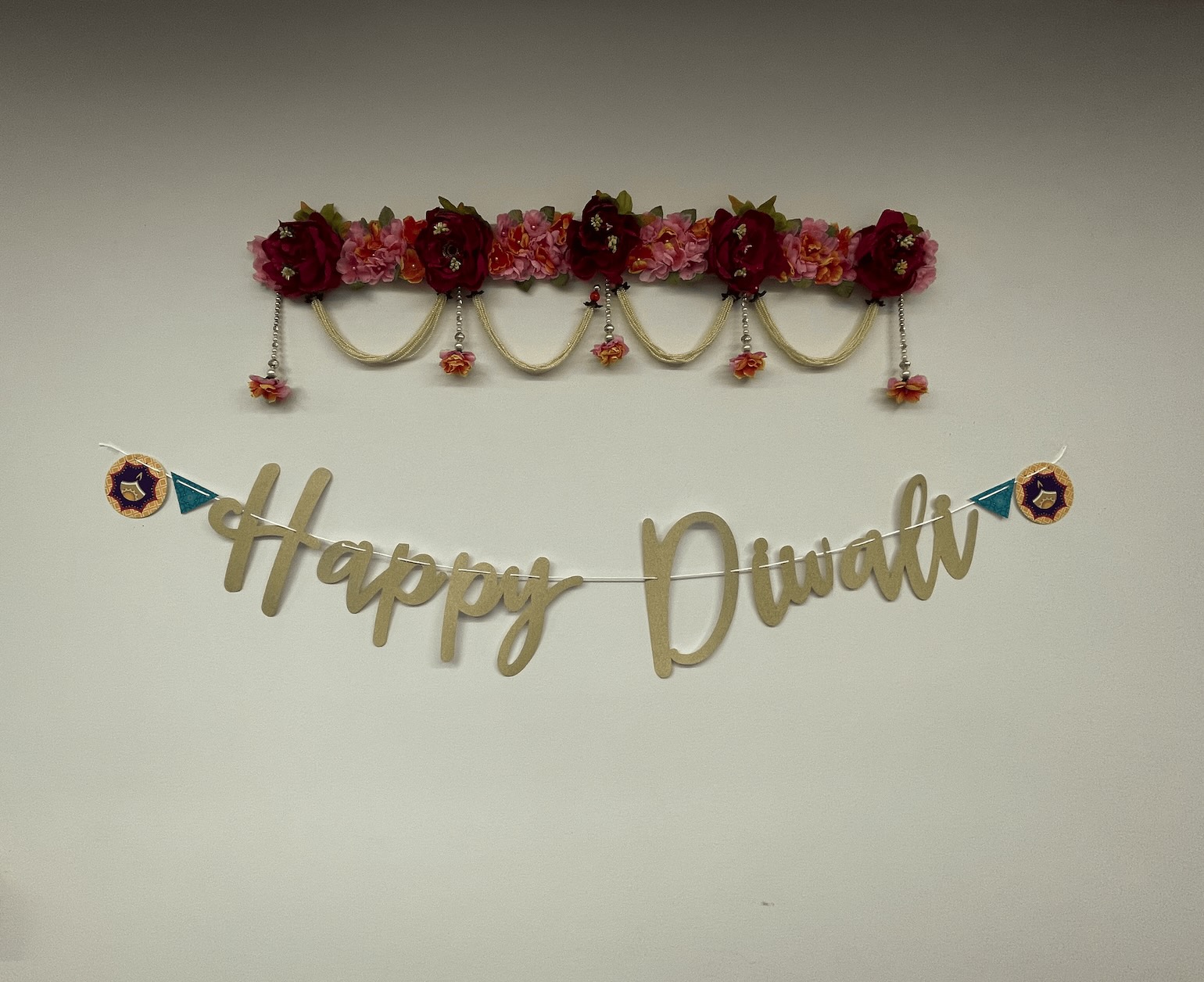 A decorative "happy Diwali" sign with letters in cursive, adorned with colorful flowers and hanging ornaments against a plain wall at GKG’s Headquarters.