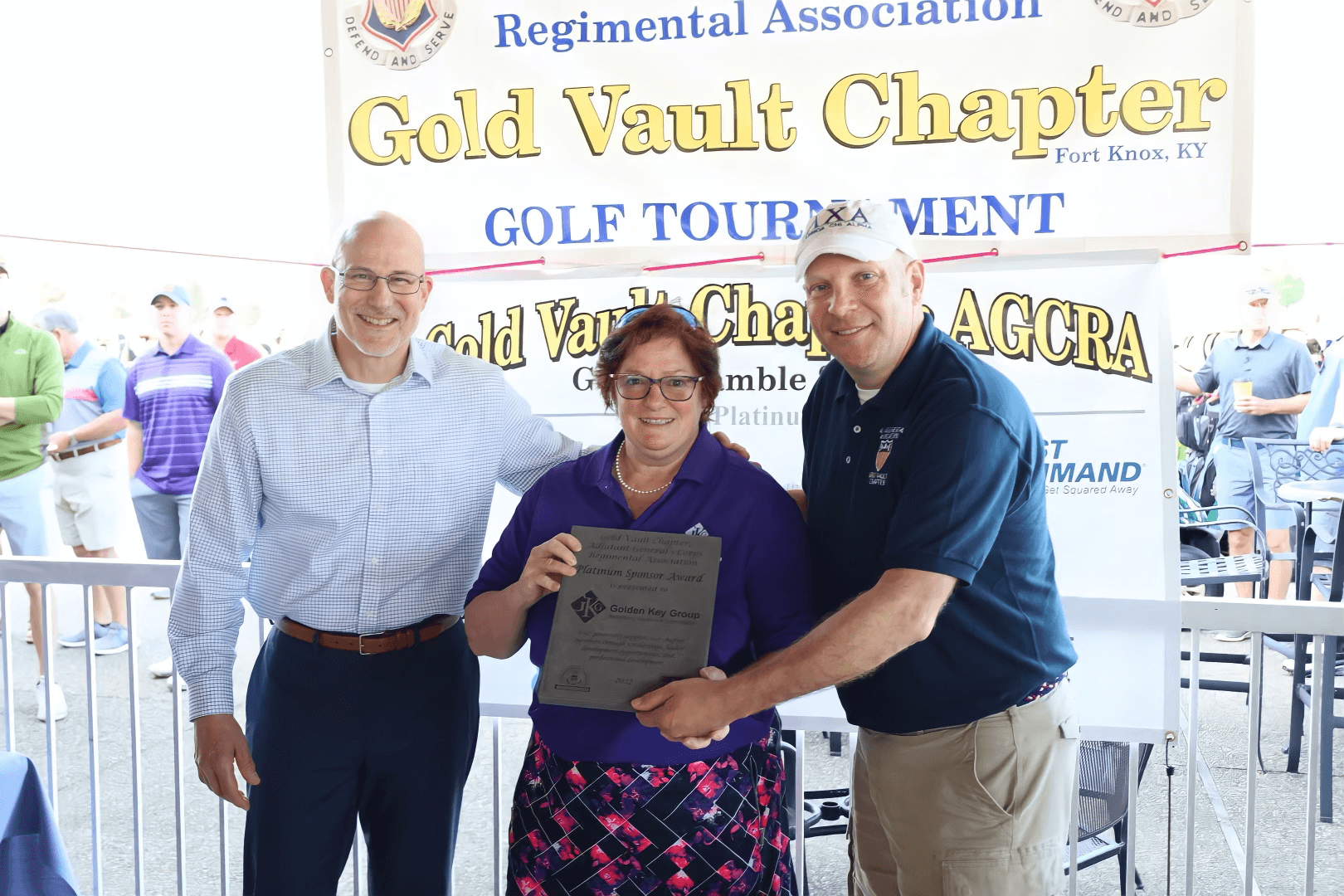 Three people pose with a certificate at the Human Capital Management chapter golf tournament in Fort Knox, KY, with banners and logos in the background.