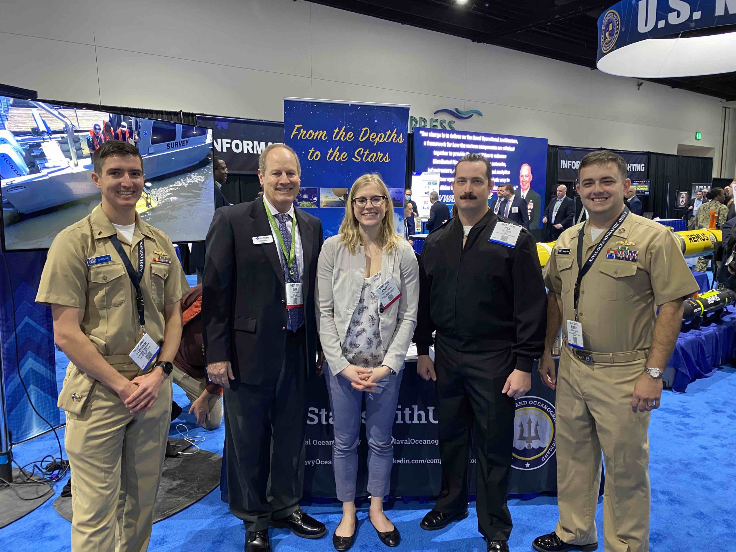 Five people posing at a Human Resources Operations exhibition booth with informational banners and a TV screen displaying a submarine.