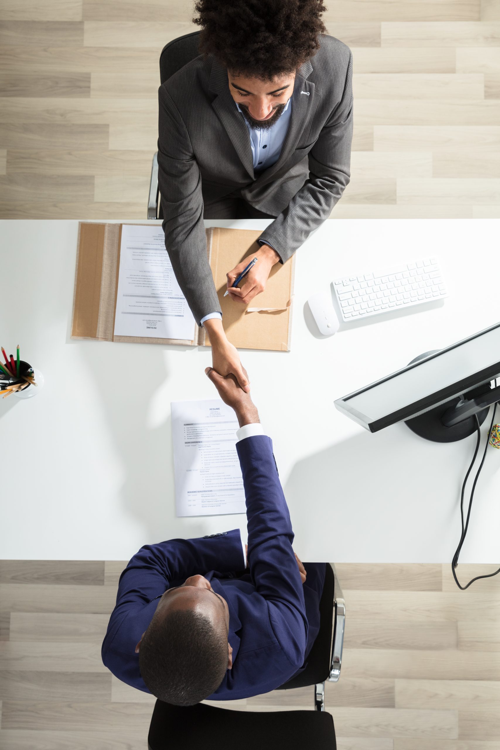 Two professional consulting services businessmen shaking hands over a desk, one standing and the other sitting, with documents in between them. View from above.