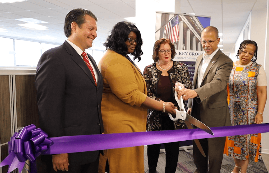 Five people participate in a ribbon-cutting ceremony with a large pair of scissors, standing behind a purple ribbon in an office setting to inaugurate the new Human Resources department.