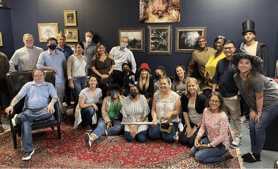 A diverse group of Human Capital Management professionals posing for a photo in a room with a red carpet and eclectic wall art, some seated and others standing.