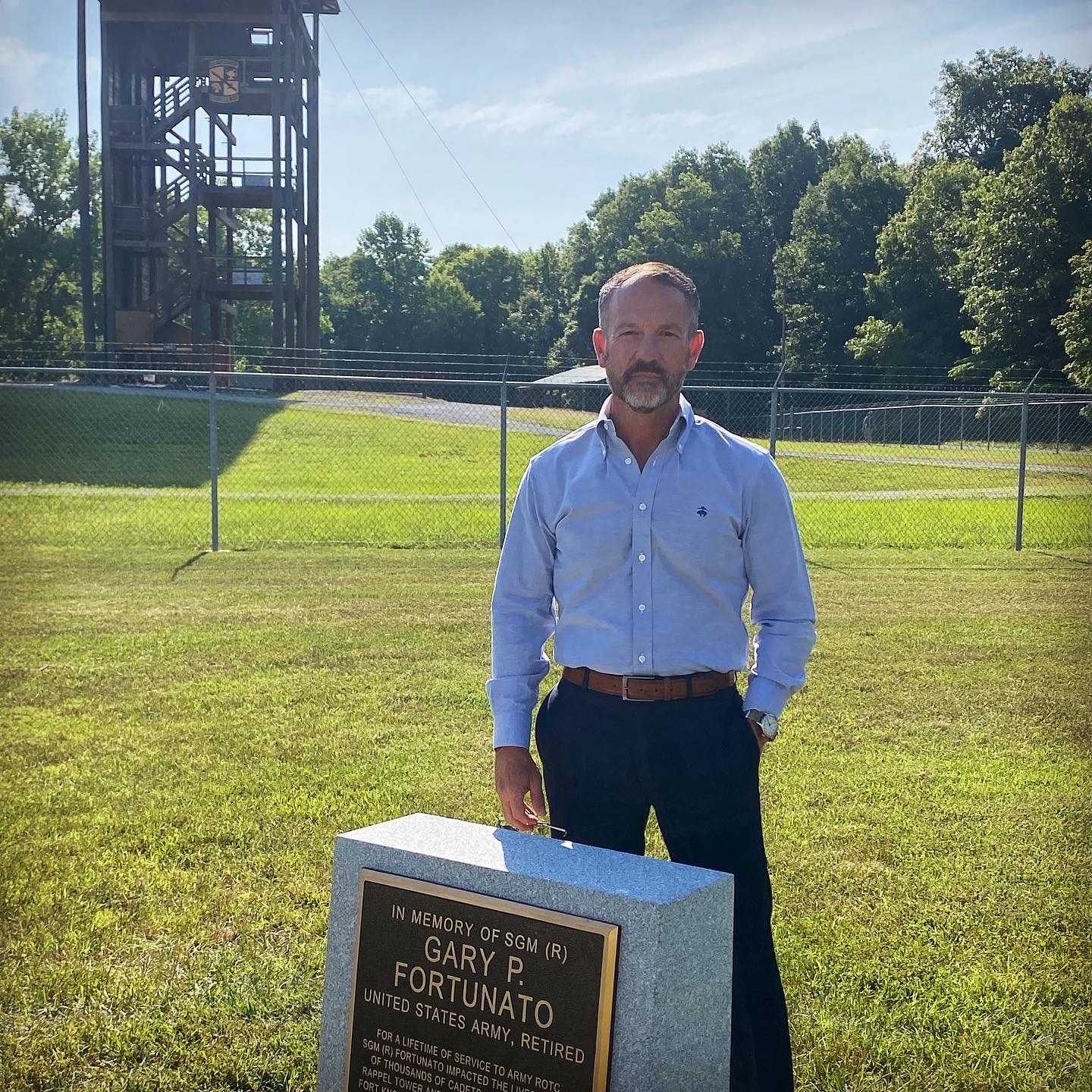 Man in a blue shirt and jeans standing next to a memorial stone in a grassy field with a tower in the background, symbolizing the commitment of Human Resources Operations to workforce optimization and honoring legacy.