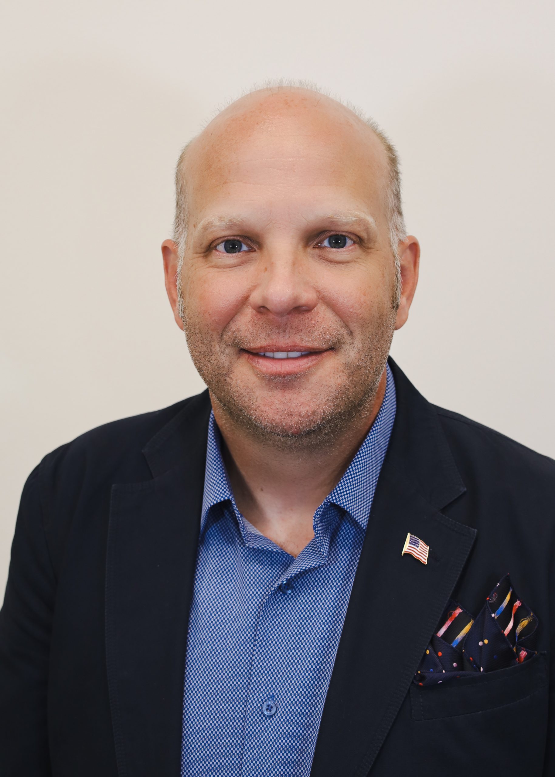 Portrait of a bald man in a navy blazer, blue shirt, and pocket square, smiling against a cream background, embodying the essence of Professional Consulting Services.